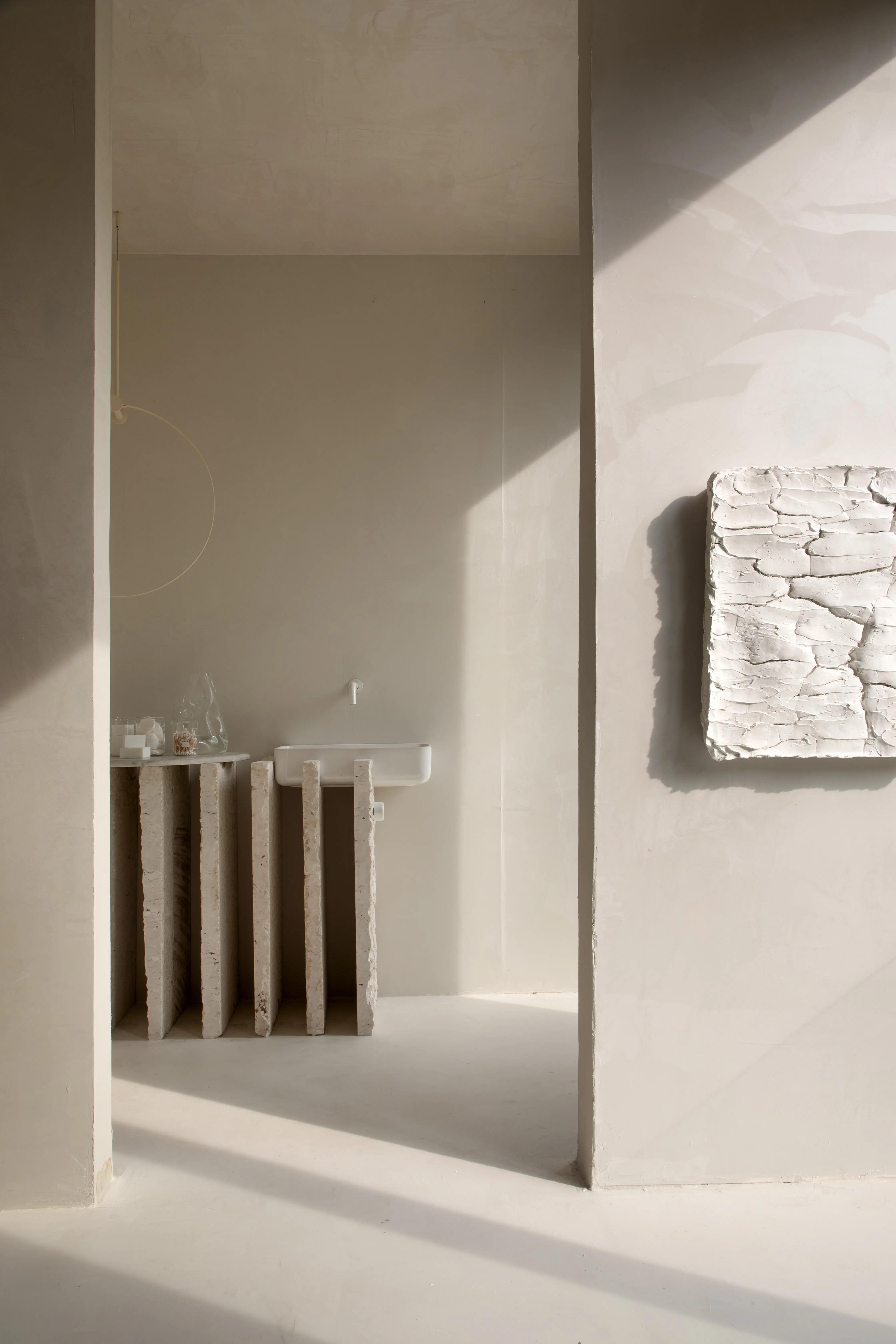 Ambiente minimalista em tons de bege, com uma pia branca fixada na parede e uma bancada de pedras verticais rústicas. À direita, uma escultura de textura orgânica em relevo está pendurada na parede. A luz natural entra suavemente, projetando sombras geométricas no chão e nas paredes.
