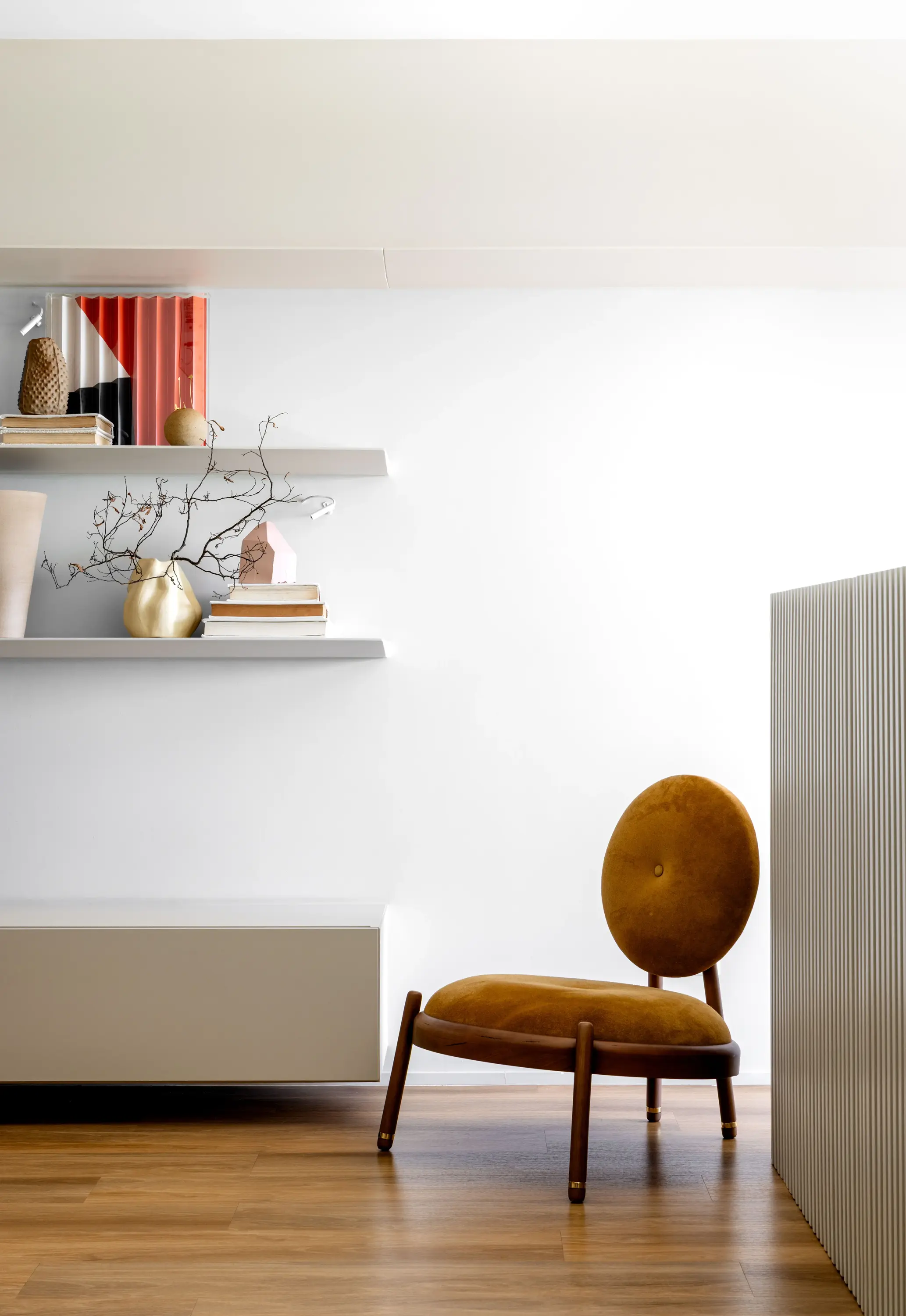 Projeto Apto Maré do Studio Guilherme Garcia. Cadeira baixa com assento e encosto arredondados em veludo mostarda, posicionada em ambiente minimalista com prateleiras decoradas com livros, esculturas e vasos sobre piso de madeira clara.