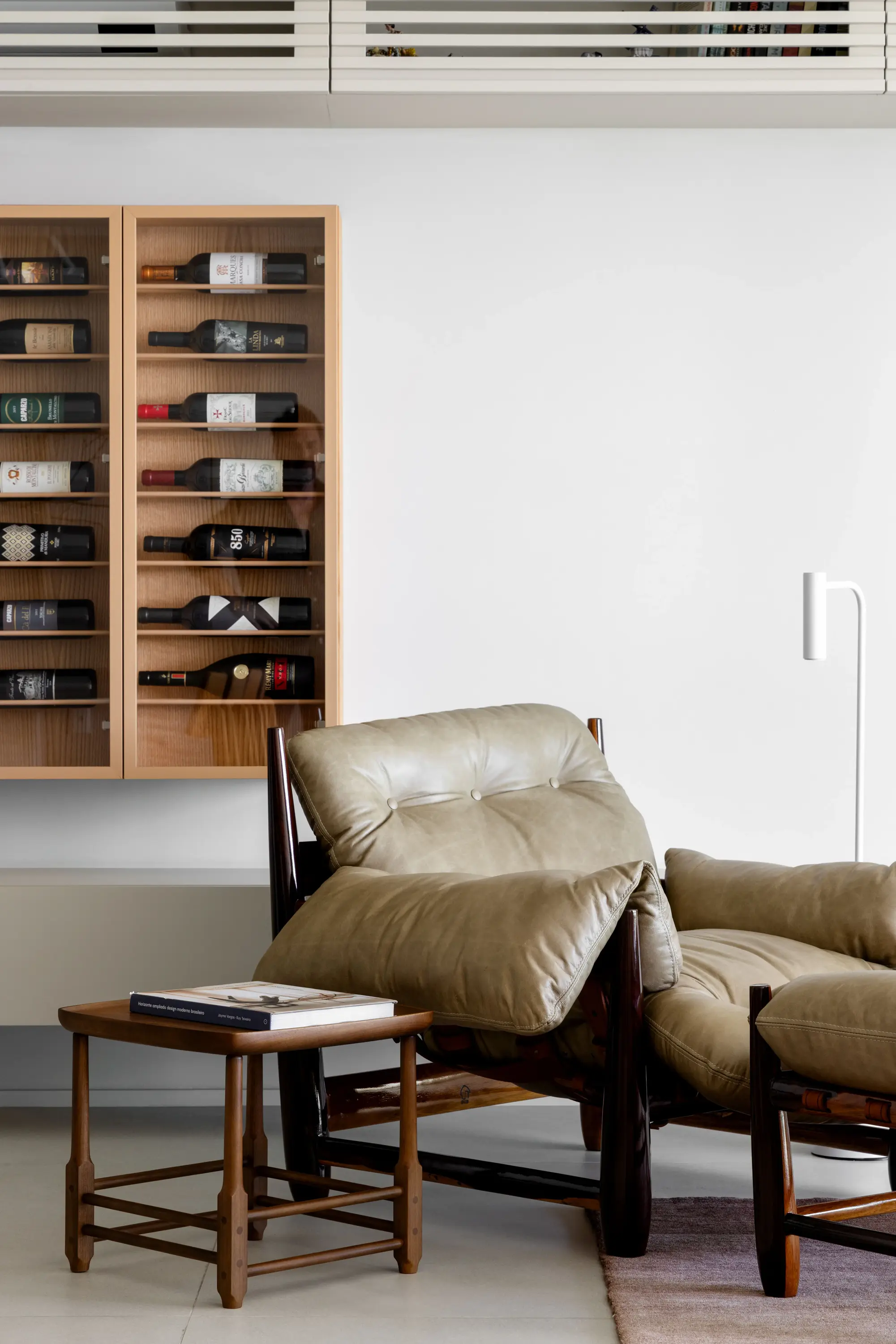 Projeto Apto Maré do Studio Guilherme Garcia. Poltrona reclinável de couro bege com pufe, ao lado de uma mesa lateral com livro, em frente a um armário de parede com garrafas de vinho organizadas em prateleiras de madeira.