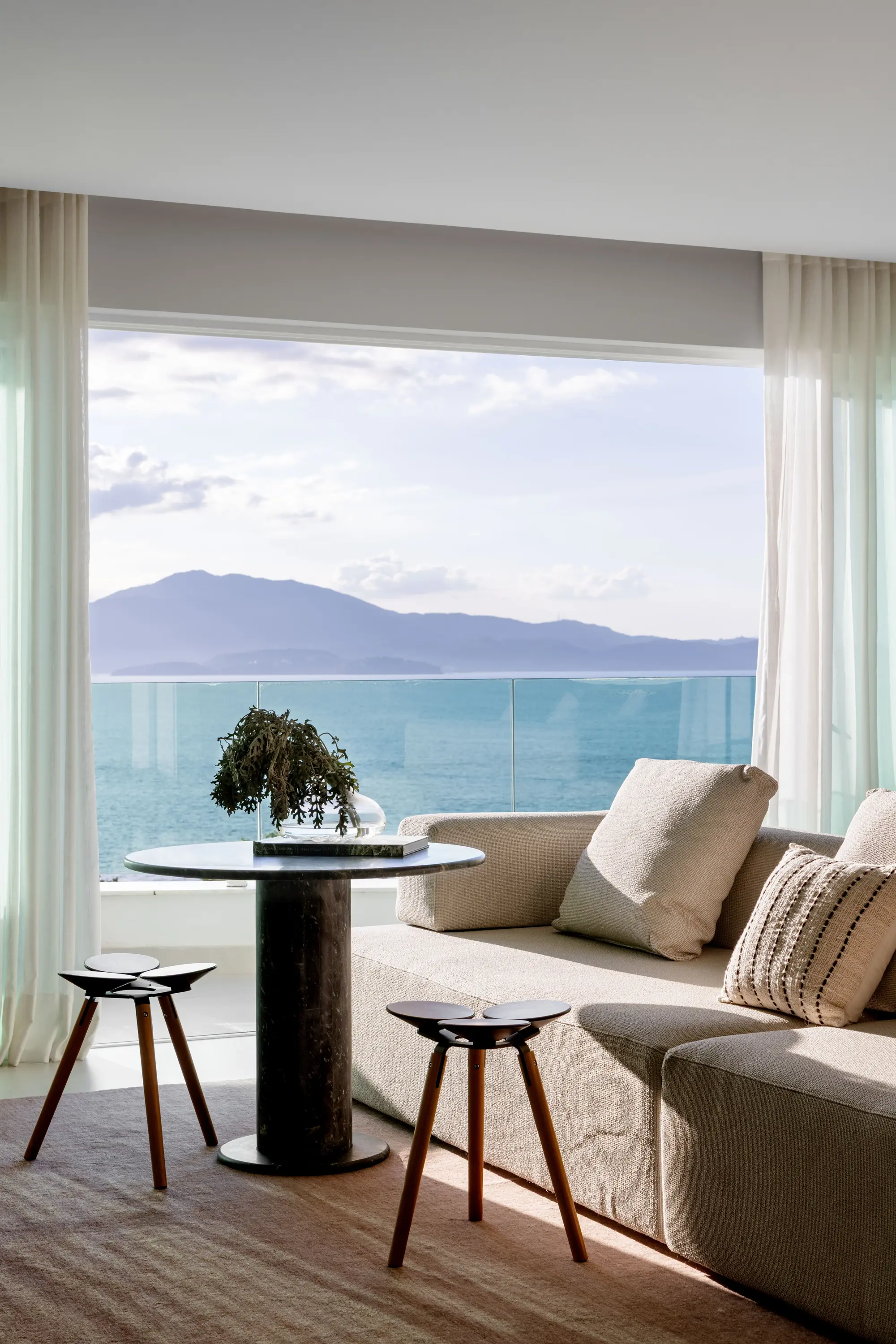 Projeto Apto Maré do Studio Guilherme Garcia. Fachada envidraçada, sacada ampla e vista para o mar em dia ensolarado. Sofá, mesa com bancos.