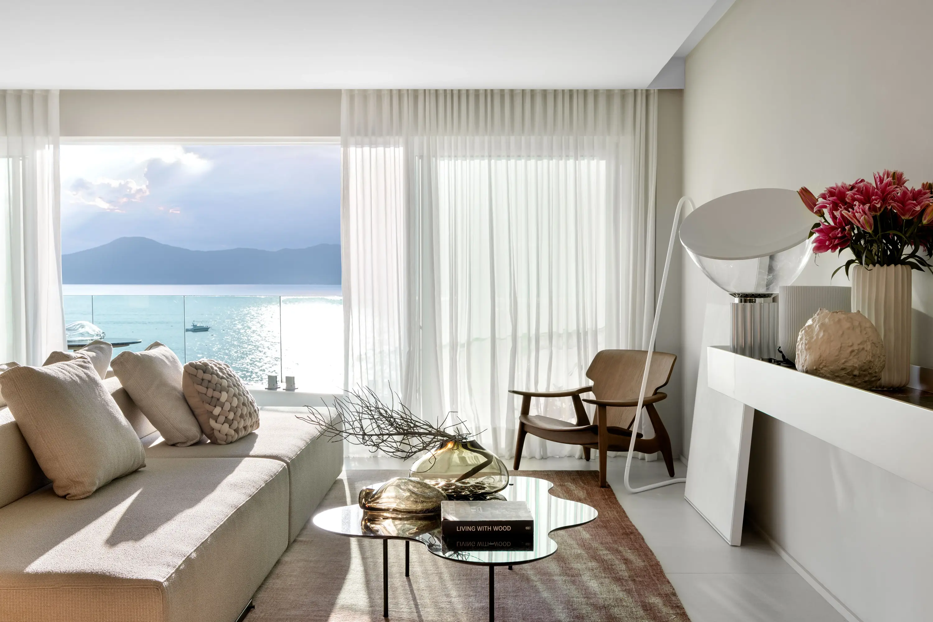 Projeto Maré do Studio Guilherme Garcia. Sala moderna com sofá, poltrona e mesa de centro, com grandes janelas que enquadram vista para o mar.
