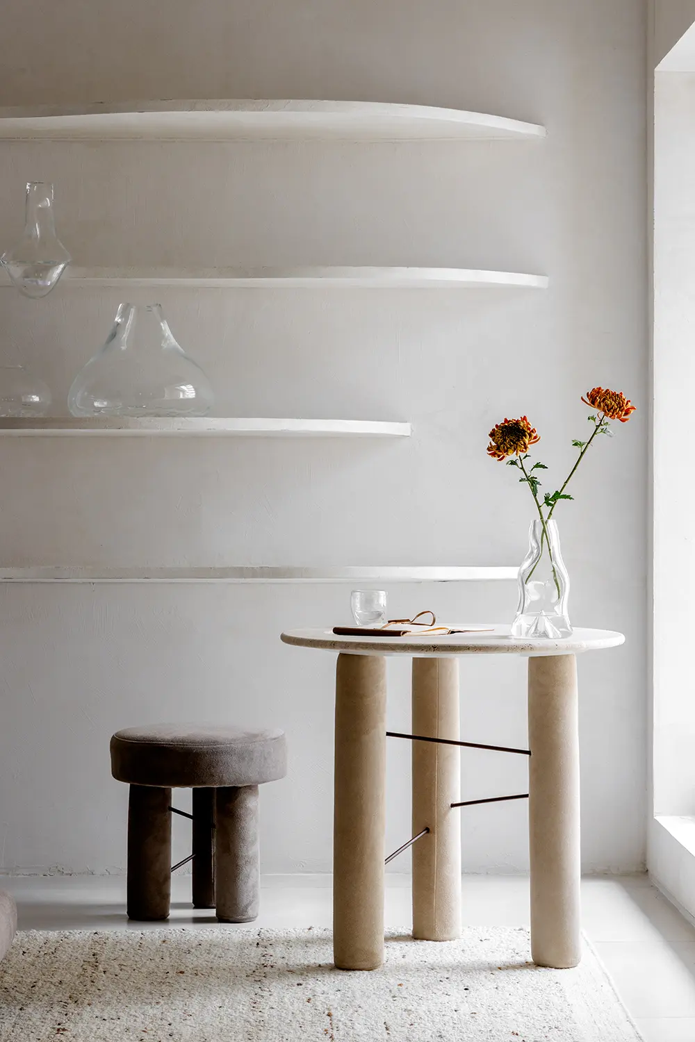 Mesa com tampo redondo e base cilíndrica ao lado de banco estofado, prateleiras orgânicas e composição minimalista no Studio Guilherme Garcia