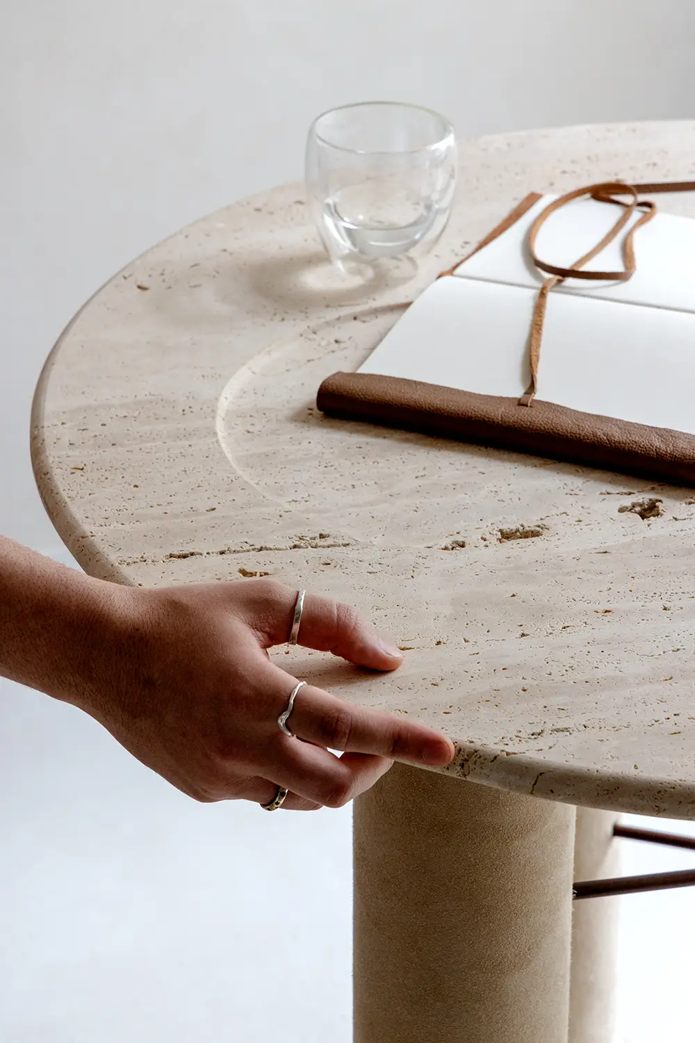 Mão tocando a borda de uma mesa redonda de pedra travertino, com um caderno de capa de couro e uma xícara de vidro transparente sobre a superfície. Coleção Perene, design funcional por Studio Guilherme Garcia