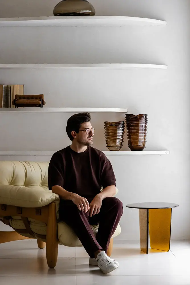 Guilherme Garcia sentado em sofá de design orgânico com prateleiras esculturais ao fundo e mesa lateral em vidro âmbar no Studio Guilherme Garcia