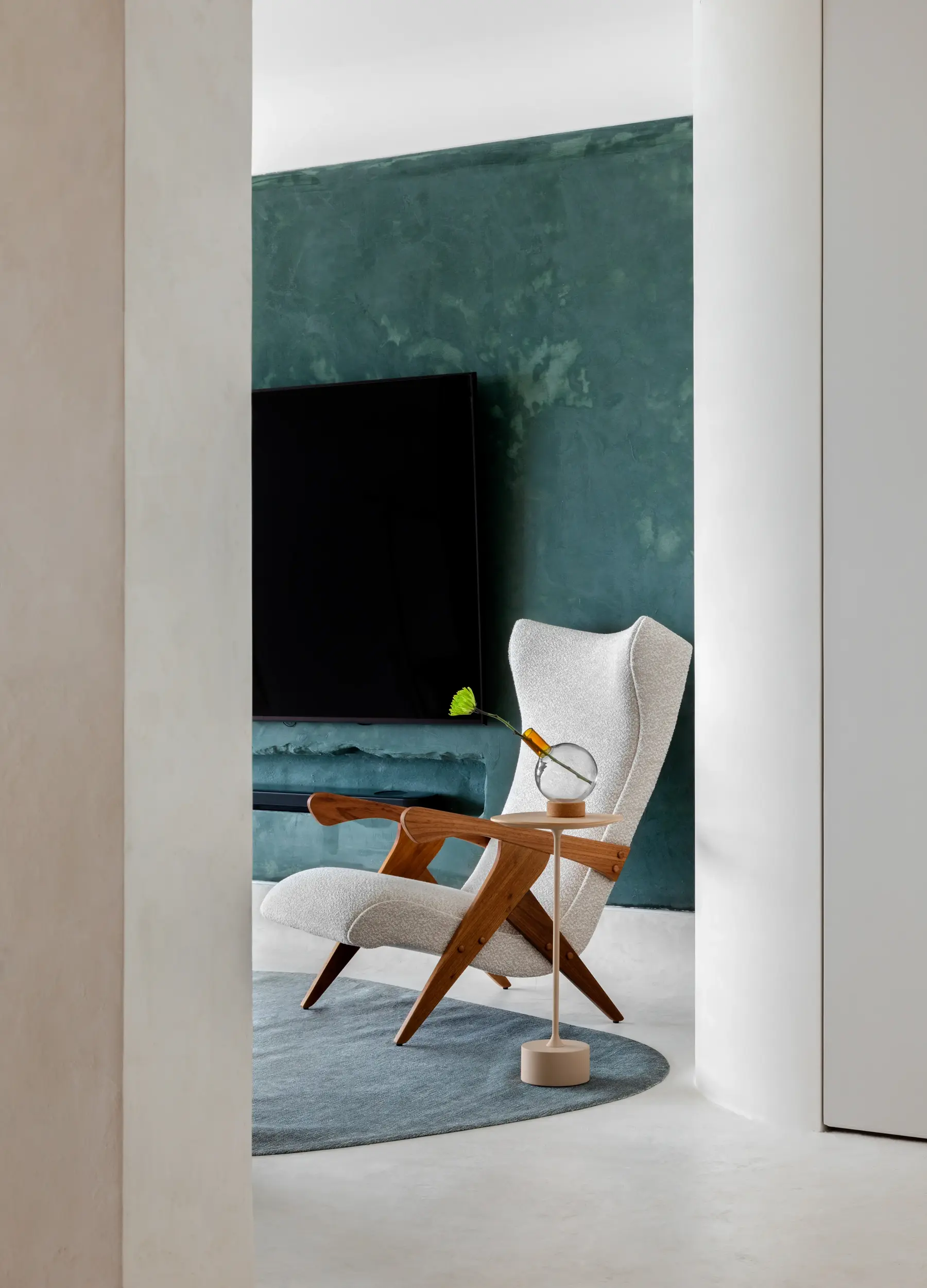 Poltrona de design com estrutura em madeira e tecido claro no projeto Apto Puro, assinado pelo Studio Guilherme Garcia, em ambiente com parede verde texturizada e mesa lateral com vaso decorativo.