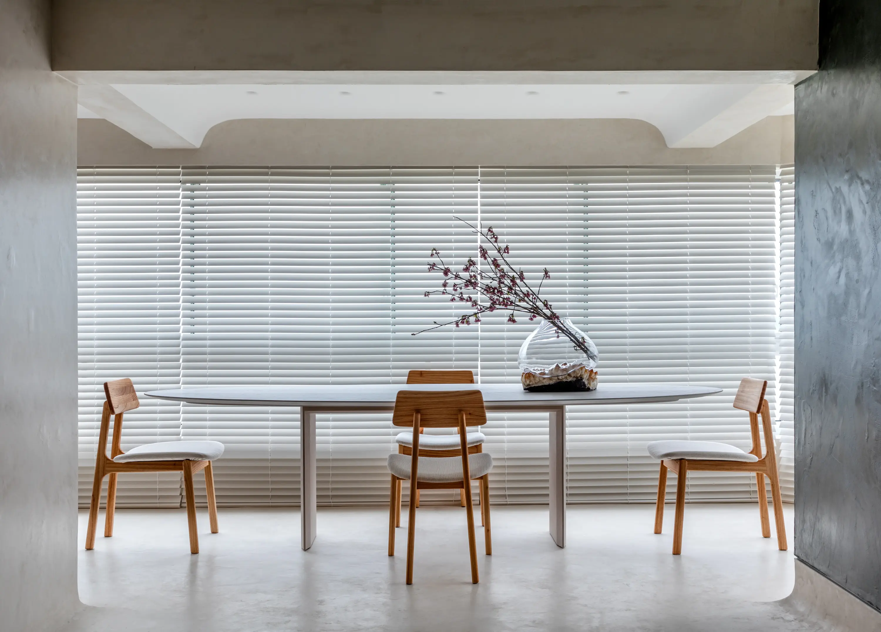 Mesa retangular branca com cadeiras de madeira clara e estofado branco, arranjo floral em vaso de vidro, ambiente minimalista com persianas brancas, projeto de interiores do Studio Guilherme Garcia