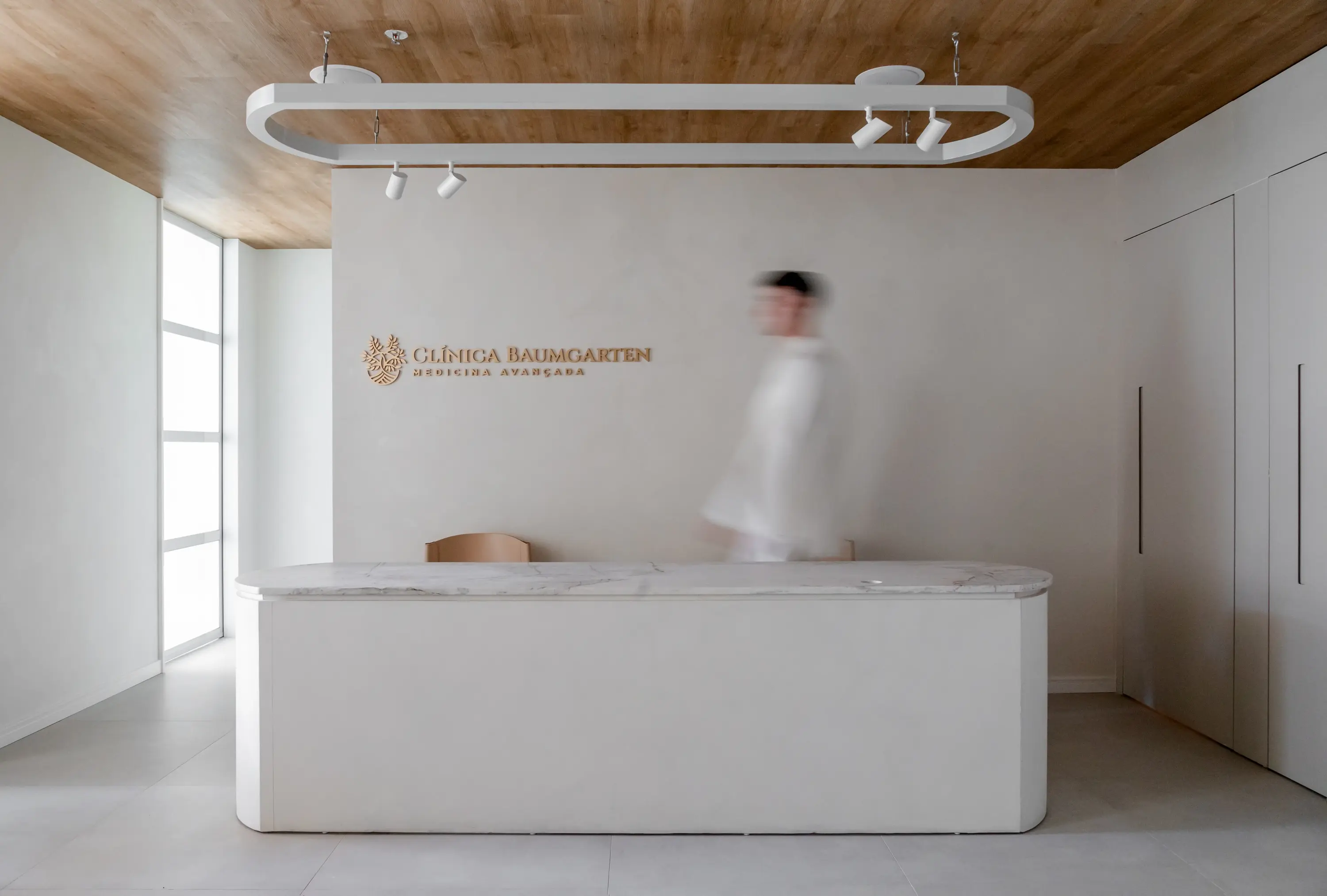 Recepção minimalista com balcão em pedra e madeira clara na Clínica Baumgarten — projeto do Studio Guilherme Garcia
